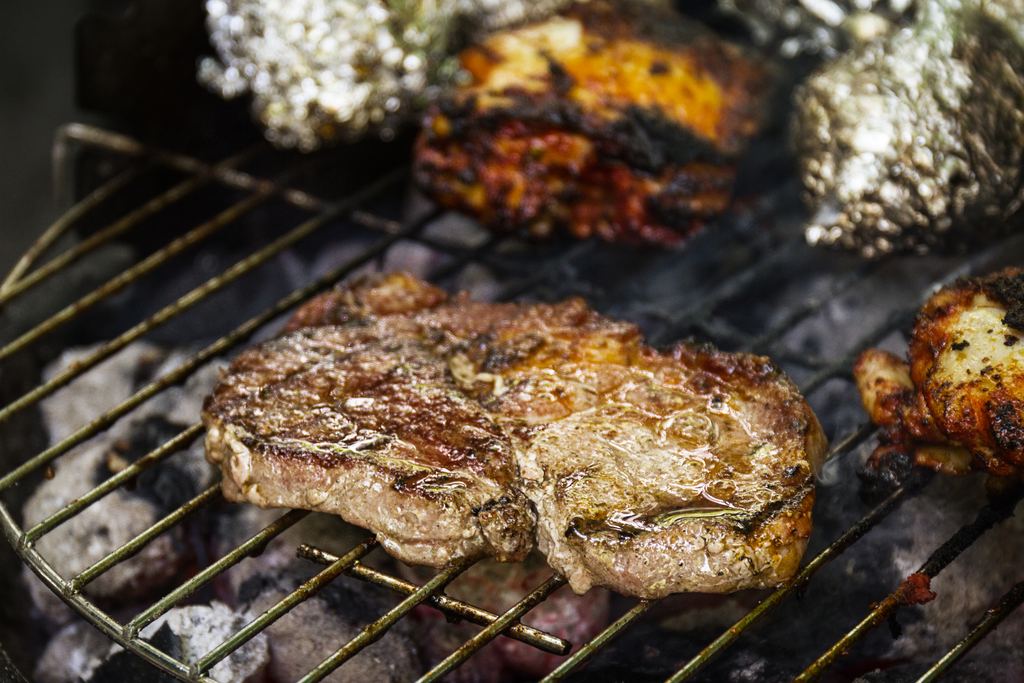 A thick cut of beef browning over an open charcoal fire on a metal grill grate
