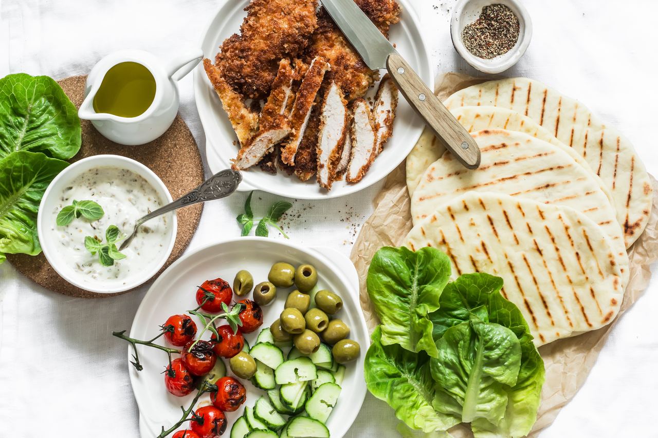 Ingredients laid out for cooking Greek gyros: pita, tzatziki, tomatoes, olives and grilled meat