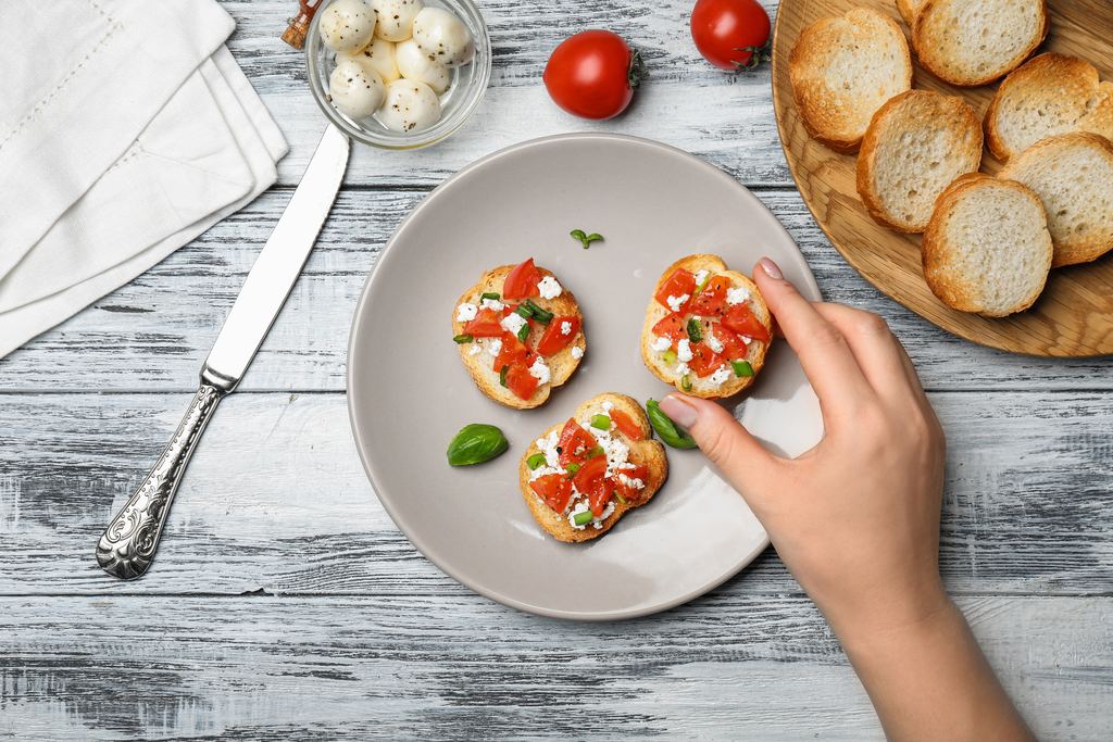 Overhead close-up of a hand picking up one small tomato bruschetta from a plate, with bread and fresh tomatoes alongside.
