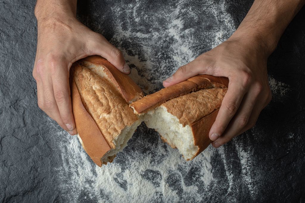 Two hands tearing a freshly baked loaf of bread in half over a dark floured surface, seen from above
