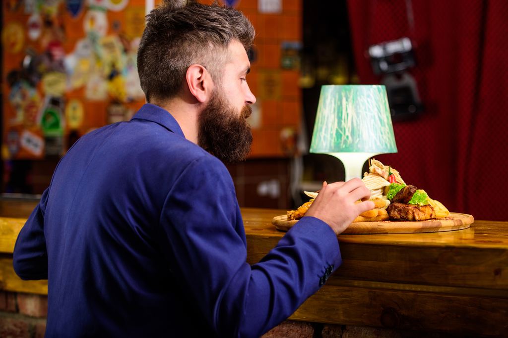 A bearded man sitting alone at a wooden bar counter with a board of fried food set in front of him.
