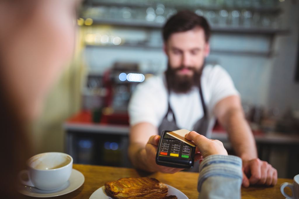 A customer holds a bank card to a payment terminal held out by a member of staff across a cafe counter