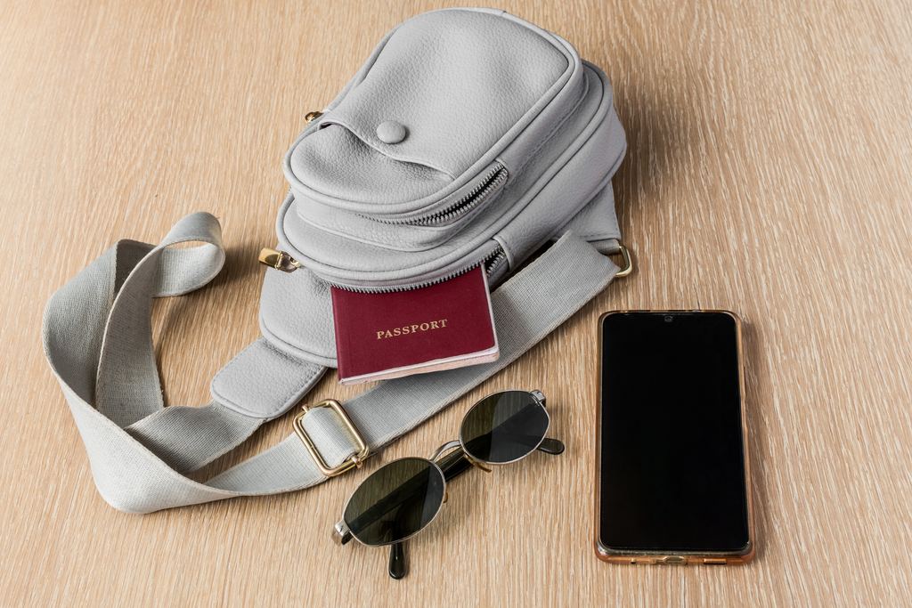 A small grey crossbody bag laid on a wooden surface with a passport, sunglasses and a smartphone beside it.