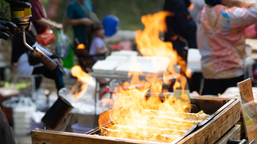 Flames rising from a charcoal grill at a street food stall, with customers waiting in the background