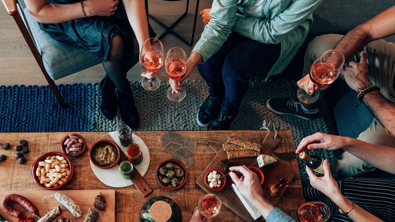 Overhead view of a table covered with small plates of cured meats, cheese and olives, surrounded by several hands and wine glasses.