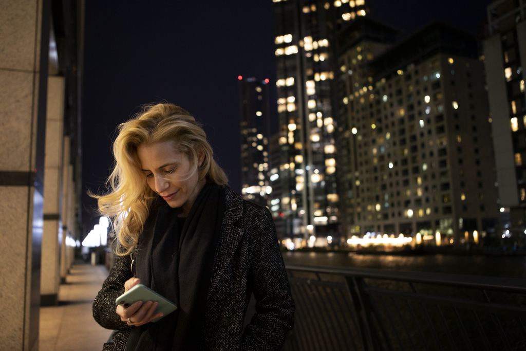 A woman standing on a city pavement in the evening looking at her smartphone, with lit buildings blurred behind her.