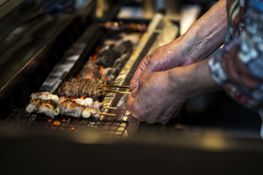 Close-up of a street food vendor's hands turning meat skewers over glowing embers on a long charcoal grill.