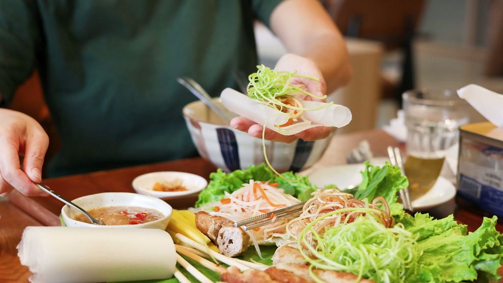 Hands rolling a fresh Vietnamese spring roll with rice paper and vegetables laid out on a table