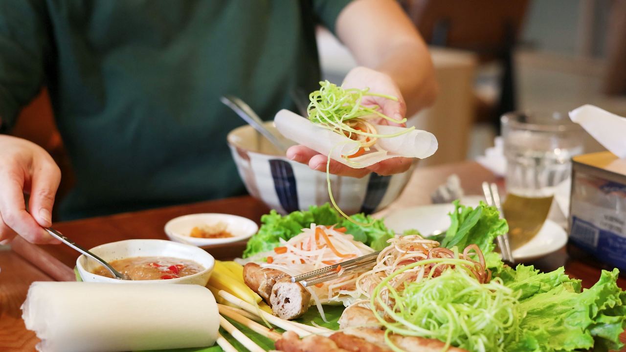 Hands rolling a fresh Vietnamese spring roll with rice paper and vegetables laid out on the table