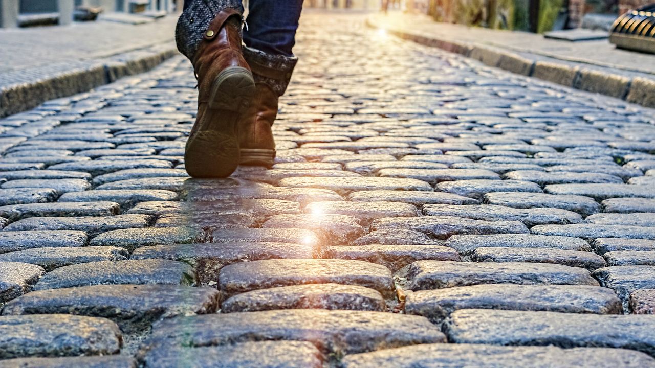 Low-angle view of a person in flat leather boots walking away down a wet cobblestone street lit by low evening sun.