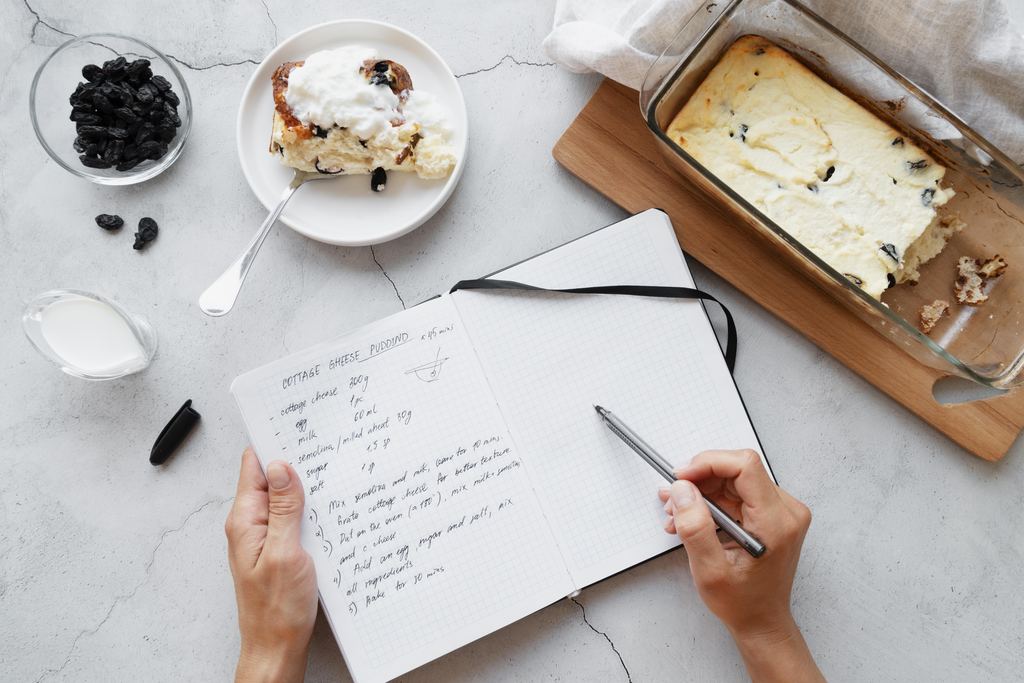 A hand writing cooking notes by hand in an open notebook, surrounded by baking ingredients on a pale kitchen surface